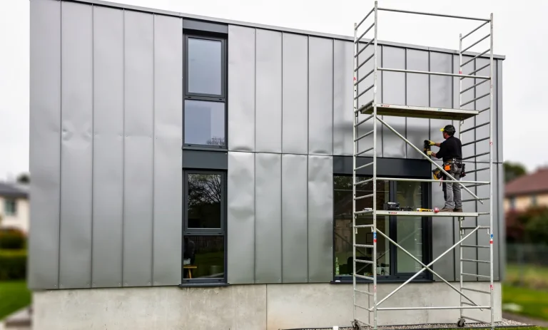 Façade d’une maison moderne en zinc où un artisan rénove une partie depuis un échafaudage sous une lumière douce.